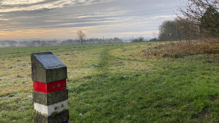An open field in winter. In the foreground a wooden post with three coloured bands marked on it.